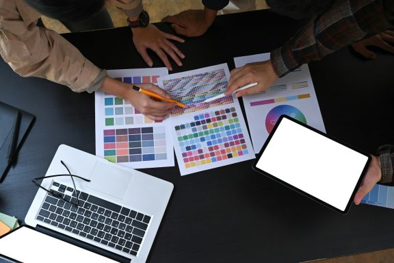 Main d'une personne tenant un crayon sur des documents colorés et un ordinateur portable.